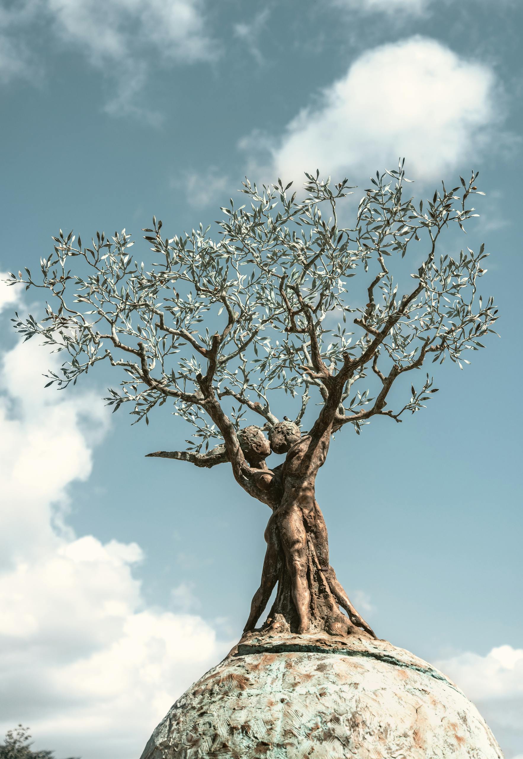 Sculpture of intertwined trees atop a globe against a blue sky, symbolizing unity.