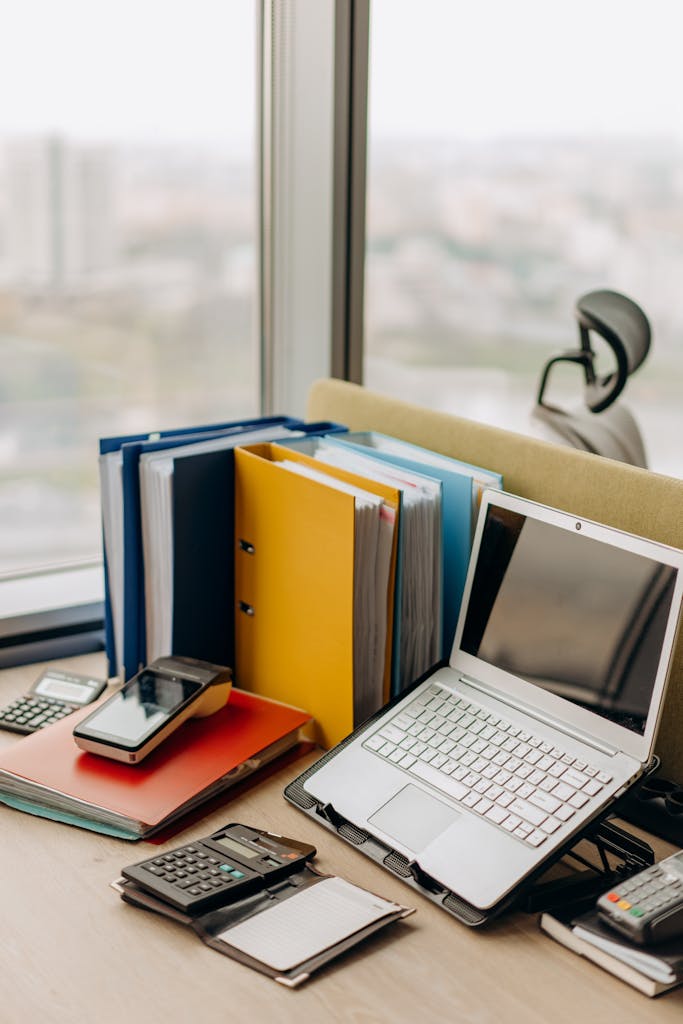 Modern workplace setup with laptop, folders, and gadgets by a window view.